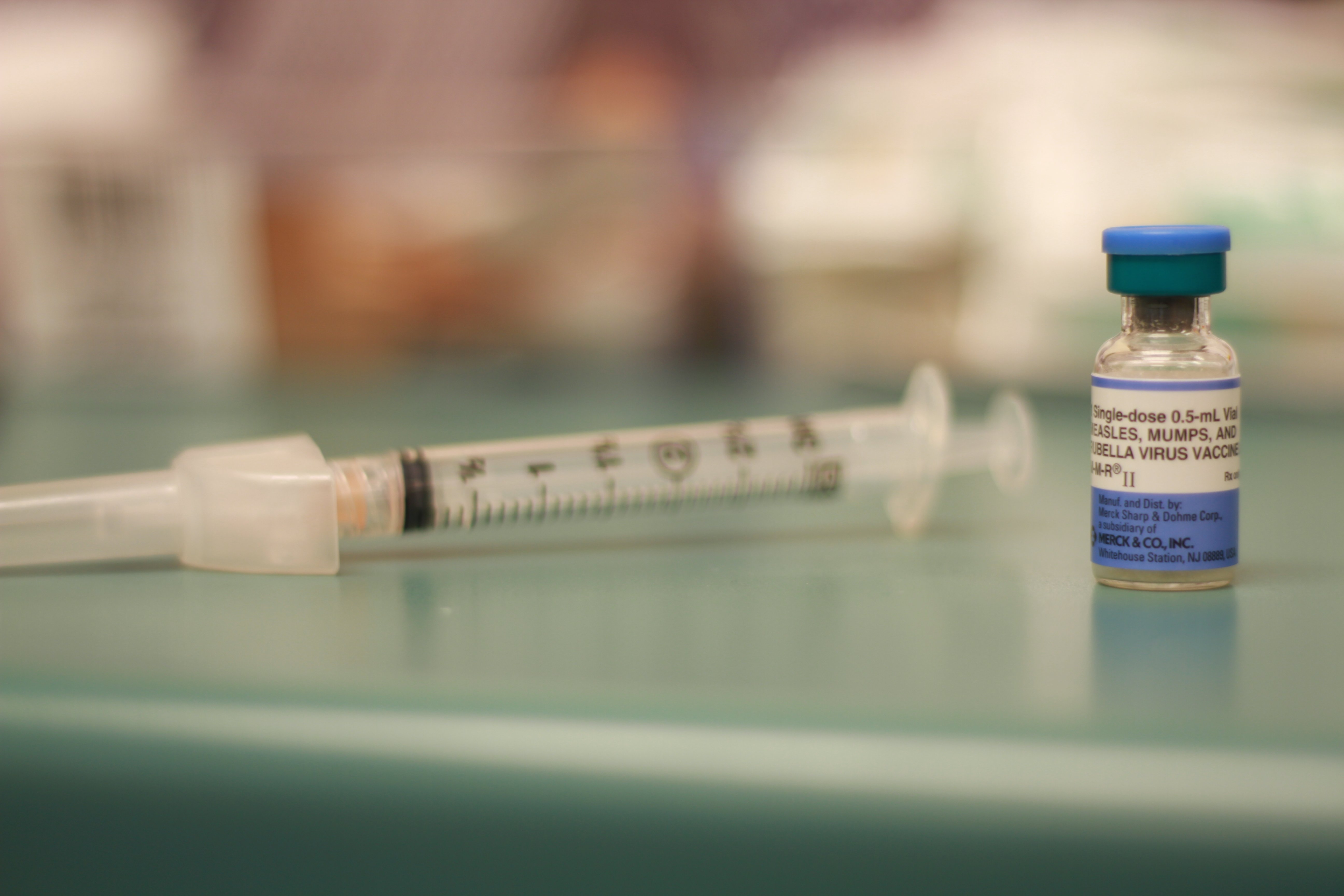 A close-up photograph of an MMR (measles, mumps and rubella) vaccine vial with a syringe in background.