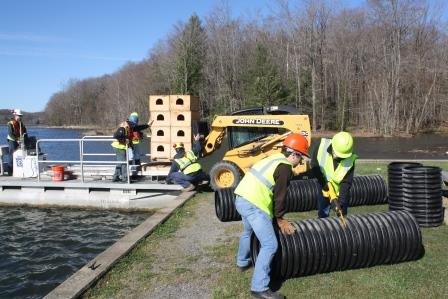 In front, left to right, Penn State DuBois Wildlife Technology Students Alec Baker and Teanna Kobuck cut section of corrugated pipe to be used in the construction of catfish spawning boxes, as other sections of the artificial habitat are loaded onto a pontoon boat behind them.  (Provided photo)
