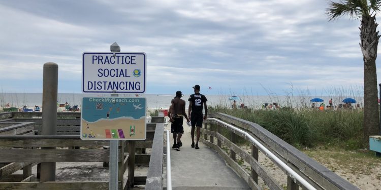 A sign in Myrtle Beach, S.C., Thursday, June 18, 2020, asks people to maintain social distancing on the beach. People are flocking to South Carolina's beaches for vacation after being cooped up by COVID-19 for months. But the virus is taking no vacation as the state has rocketed into the top five in the country in cases divided by population.   Jeffrey Collins / AP Photo