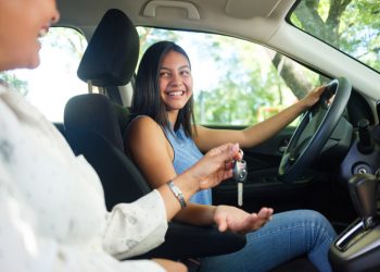 A side view photo of a teenage girl in a car as her mother sits next to her and hands her the keys to drive.