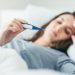 Woman with flu virus lying in bed, she is measuring her temperature with a thermometer and touching her forehead