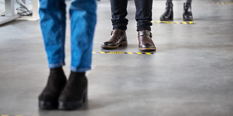 Low angle shot of business people standing behind social distancing signage on office floor. People in queue at office for safe entry checks post corona virus pandemic.