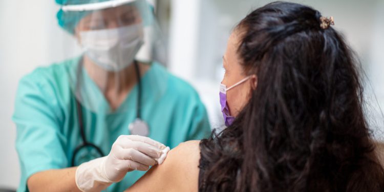 Female doctor in protective workwear disinfecting female patient's skin for vaccination; prevention and immunization from corona virus infection