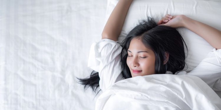 Beautiful young pretty Asian woman wake up and make happy smile with white shirt at the white bed in the morning.