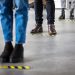 Low angle shot of business people standing behind social distancing signage on office floor. People in queue at office for safe entry checks post corona virus pandemic.