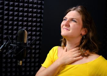 A young woman holding her throat near the microphone, black background. The voice of the singer when recording a song in the music Studio. Voice failure, hoarseness, pain in the vocal cords.