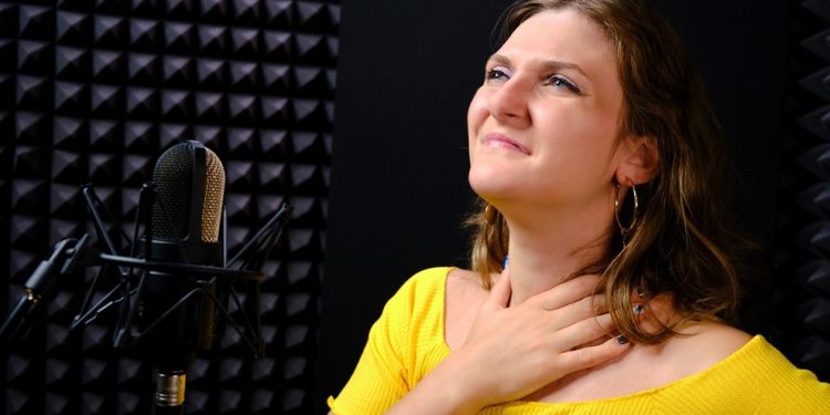 A young woman holding her throat near the microphone, black background. The voice of the singer when recording a song in the music Studio. Voice failure, hoarseness, pain in the vocal cords.