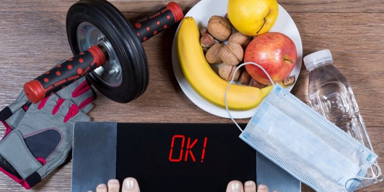 Girl checks her weight after quarantine. Digital scales with word ok surrounded by sport accessories, healthy food, water bottle and face mask. Concept of healthy lifestyle during self-isolation.