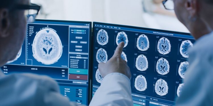 In Control Room Doctor and Radiologist Discuss Diagnosis while Watching Procedure and Monitors Showing Brain Scans Results, In the Background Patient Undergoes MRI or CT Scan Procedure.