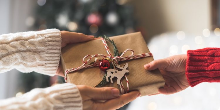 Woman in sweater giving a wrapped Christmas gift box to child. Glowing snow bokeh, fir tree. Winter holidays.