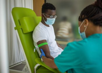 Young man on blood donation at medical clinic, nurse and him are wearing protective face masks for protection against coronavirus
