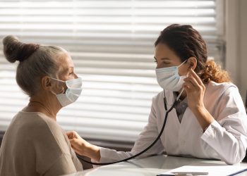Caring young female doctor in medical facemask hold stethoscope listen to elderly patient heart in hospital. Woman GP or physician do regular checkup examine mature client at consultation in clinic.
