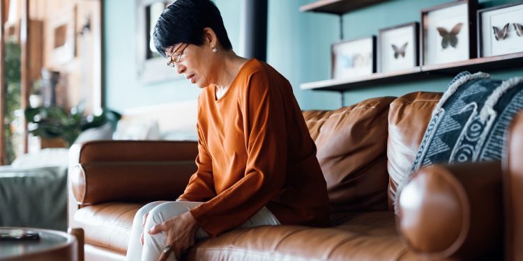 A distraught senior Asian woman feeling unwell, suffering from pain in leg while sitting on sofa in the living room at home