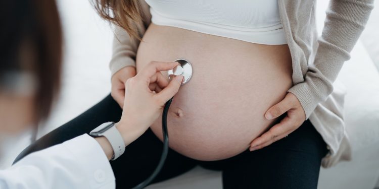 Close up of a female doctor doing checkup on an Asian pregnant woman, examining the belly with stethoscope. Prenatal exam. Pregnancy health and wellbeing concept