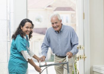 A home healthcare worker helping a senior man with a walker walking through the front door of his house. The nurse is a mature woman, a Pacific Islander, wearing scrubs. They are looking up at the camera, smiling.