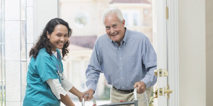 A home healthcare worker helping a senior man with a walker walking through the front door of his house. The nurse is a mature woman, a Pacific Islander, wearing scrubs. They are looking up at the camera, smiling.