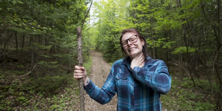 Young woman winces in discomfort as she is slapping her neck to kill a bug that is biting her while she is on a beautiful forest hike. She is wearing a blue plaid shirt, and eyeglasses and holding a walking stick while standing on a trail between tall trees.
