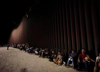 Migrants wait along a border wall Aug. 23, 2022, after crossing from Mexico near Yuma, Ariz. U.S. immigration offices have become so overwhelmed with processing migrants for court that some some asylum-seekers who crossed the border at Mexico may be waiting a decade before they even get a date to see a judge.

Gregory Bull / AP Photo