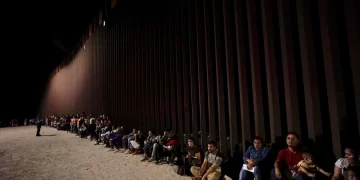 Migrants wait along a border wall Aug. 23, 2022, after crossing from Mexico near Yuma, Ariz. U.S. immigration offices have become so overwhelmed with processing migrants for court that some some asylum-seekers who crossed the border at Mexico may be waiting a decade before they even get a date to see a judge.

Gregory Bull / AP Photo