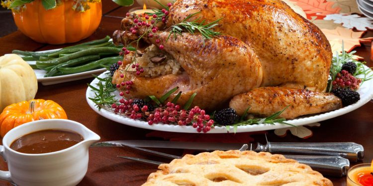 Roasted pepper turkey for Thanksgiving, garnished with pink pepper, blackberry, and fresh rosemary twigs on a dinner table decorated with mini pumpkins, beans, carrots, baked potato, pie, cranberry relish, gravy, flowers, candles, and flutes of champagne."n