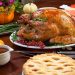 Roasted pepper turkey for Thanksgiving, garnished with pink pepper, blackberry, and fresh rosemary twigs on a dinner table decorated with mini pumpkins, beans, carrots, baked potato, pie, cranberry relish, gravy, flowers, candles, and flutes of champagne."n