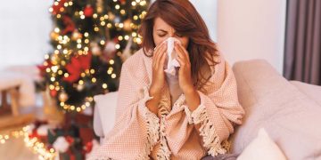 Sick woman having fever and flu, lying in bed covered with blanket blowing nose while spending Christmas day relaxing at home