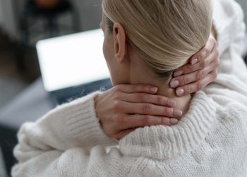 back view of female in white sweater feeling discomfort and ache, touching and massaging neck with hands near laptop in room, overwork concept