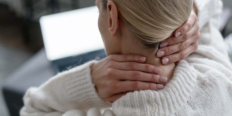 back view of female in white sweater feeling discomfort and ache, touching and massaging neck with hands near laptop in room, overwork concept