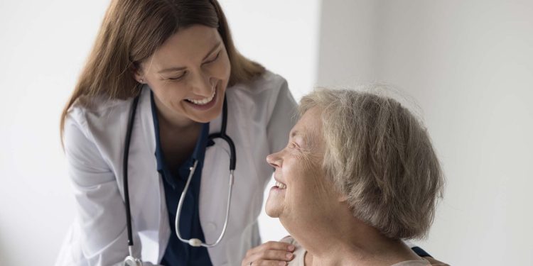 Cheerful female doctor with stethoscope touching shoulders of happy elderly senior patient woman, embracing old lady with care, support, talking, smiling, laughing