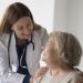 Cheerful female doctor with stethoscope touching shoulders of happy elderly senior patient woman, embracing old lady with care, support, talking, smiling, laughing