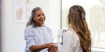 The mature woman smiles while shaking hands with the female healthcare professional as she introduces herself.
