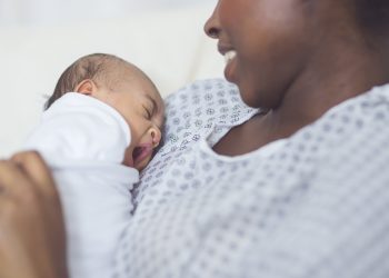 A beautiful young African American mother in a hospital gown gently holds her infant in her arms and looks at her affectionately. The swaddled baby's eyes closed as she yawns.