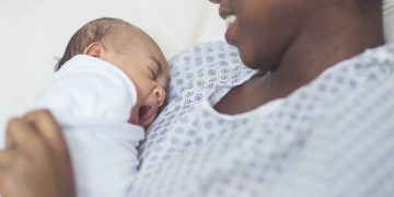 A beautiful young African American mother in a hospital gown gently holds her infant in her arms and looks at her affectionately. The swaddled baby's eyes closed as she yawns.