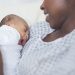 A beautiful young African American mother in a hospital gown gently holds her infant in her arms and looks at her affectionately. The swaddled baby's eyes closed as she yawns.