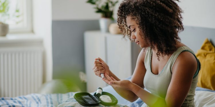 Teenage girl testing her blood sample on blood sugar meter at home. Girl holding glucometer with test strip, waiting for results. Paediatric diabetes in teenage girl and life with chronic illness.