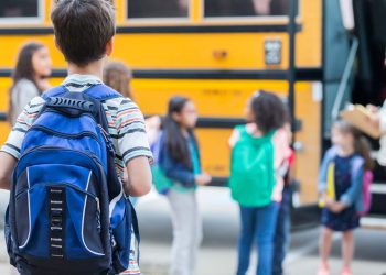 Rear view of elementary age boy waiting to get on school bus. His classmates are loading the bus in the background.