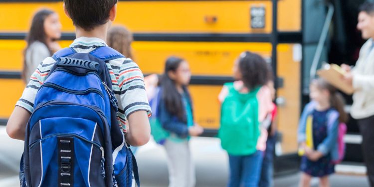 Rear view of elementary age boy waiting to get on school bus. His classmates are loading the bus in the background.