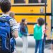 Rear view of elementary age boy waiting to get on school bus. His classmates are loading the bus in the background.