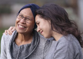 A senior woman with cancer is embraced and comforted by her adult daughter as they sit outside on a fall evening. The mother is smiling and laughing while the daughter is squeezing her mother affectionately and smiling as well.