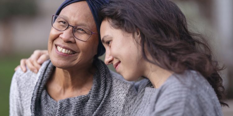 A senior woman with cancer is embraced and comforted by her adult daughter as they sit outside on a fall evening. The mother is smiling and laughing while the daughter is squeezing her mother affectionately and smiling as well.